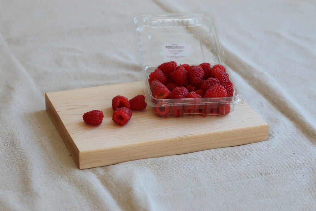 Raspberries on a wooden board with a clear plastic container of raspberries on a beige fabric background